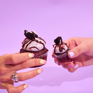 Two hands holding Vegan Red Velvet Volcano Cupcakes with Oreo and ganache topping on purple background